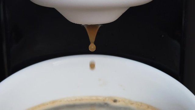 finishing of brewing espresso with a capsule coffee machine in black and white colours, last drops of coffee into a full white cup, end of brewing, side view