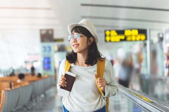 Happy Asian woman wear glasses, hat with yellow backpack is holding flying ticket, passport while waiting for the flight at the hall of airport. 