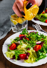 female hand squeezes lemon juice on various fresh mix salad leaves with tomato in white plate on wooden background.