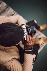 Fototapeta premium Close-up shot of young male photographer in brown coat taking pictures with a small mirrorless camera sitting on a wooden lake house deck in lake Plansee, Austria