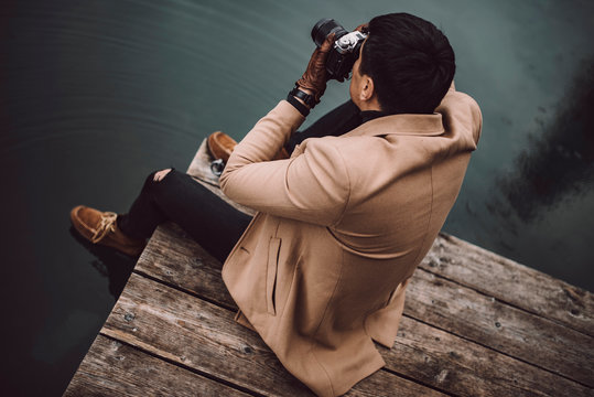 Full body shot of young male photographer in brown coat taking pictures with a small mirrorless camera while sitting on a wooden lake house deck in lake Plansee, Austria