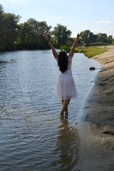 A dark haired young girl in a white dress walks barefoot along the river Bank 