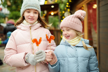 Two adorable sisters having rooster-shaped lollipops on traditional Christmas fair in Riga, Latvia. Children enjoying sweets, candies and gingerbread on Xmas market.