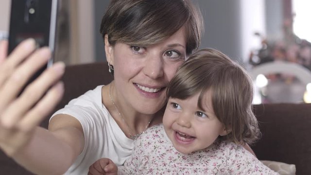 Portrait of a beautiful young mother and her little daughter taking a selfie with a smartphone at home on the couch. Shot in 6k, cine lens.	