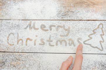 Female hand writing with your finger on the background of flour with Christmas. Christmas background with white flour and silhouette of Christmas tree with inscription. The view from the top.