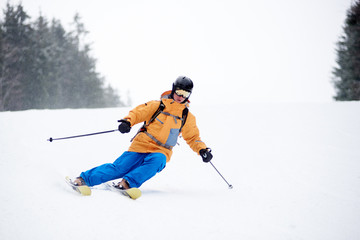 Man skier in ski sportswear, helmet and goggles, riding down the snowy slope, doing skillful maneuvers on skis. Proficient skiing in snowfall cold weather. Monochrome winter view on background.