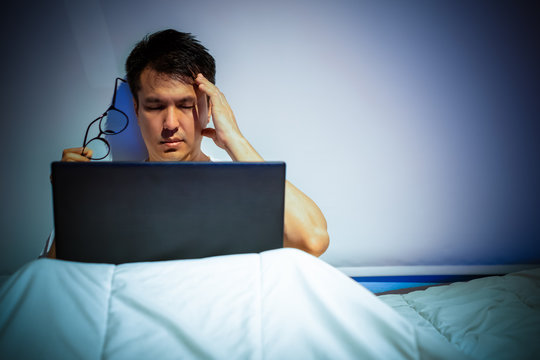 headache, tense young asian man working on laptop computer in bed at night