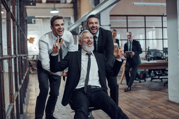 Cheerful businessmen pushing their boss on the office chair while running in the hallway