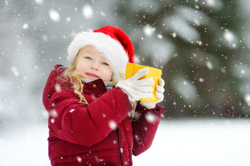 Adorable little girl wearing Santa hat holding Christmas gift on beautiful winter day.
