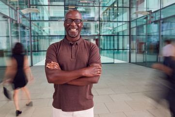 Portrait Of Mature Businessman With Crossed Arms Standing In Lobby Of Busy Modern Office