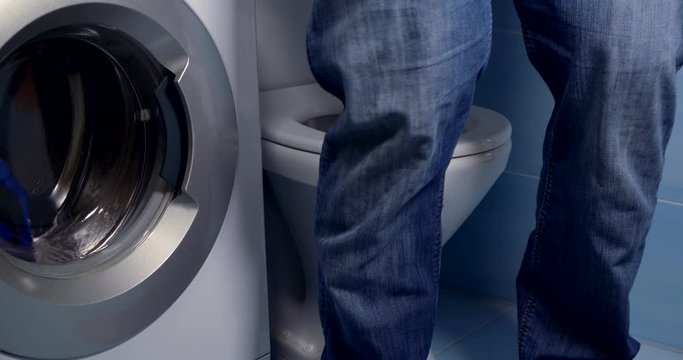 Close-up a man in a shirt lowers his jeans and underpants, sits down on the toilet, rubs his hands, next to it is a washing machine in which the clothes are washed.