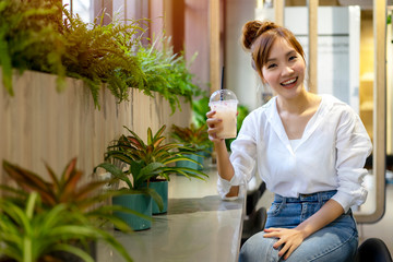 Asian young woman sitting at coworking space office during a break, holding cold beverage plastic glass in hand with smiling and cheerful face. Relaxing zone for worker with green area