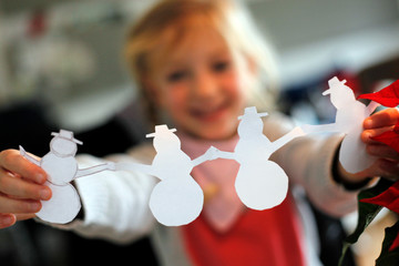 Kleines fröhliches Mädchen bastelt Girlande Motiv weißer Schneemann aus Papier im Winter in der Adventszeit an Weihnachten - obrazy, fototapety, plakaty