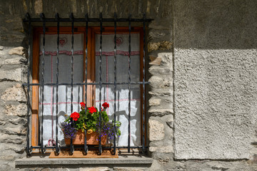 Fototapeta na wymiar Close-up of the window of a mountain stone chalet with a red flowering potted plant on the windowsill, Chianale, Cuneo, Piedmont, Italy
