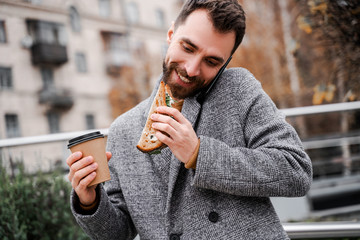Confident successful professional person eating sandwich and drink coffee take away, calling mobile phone for work. Multitasking businessman. Smile and enjoy break time. Hungry worker having lunch