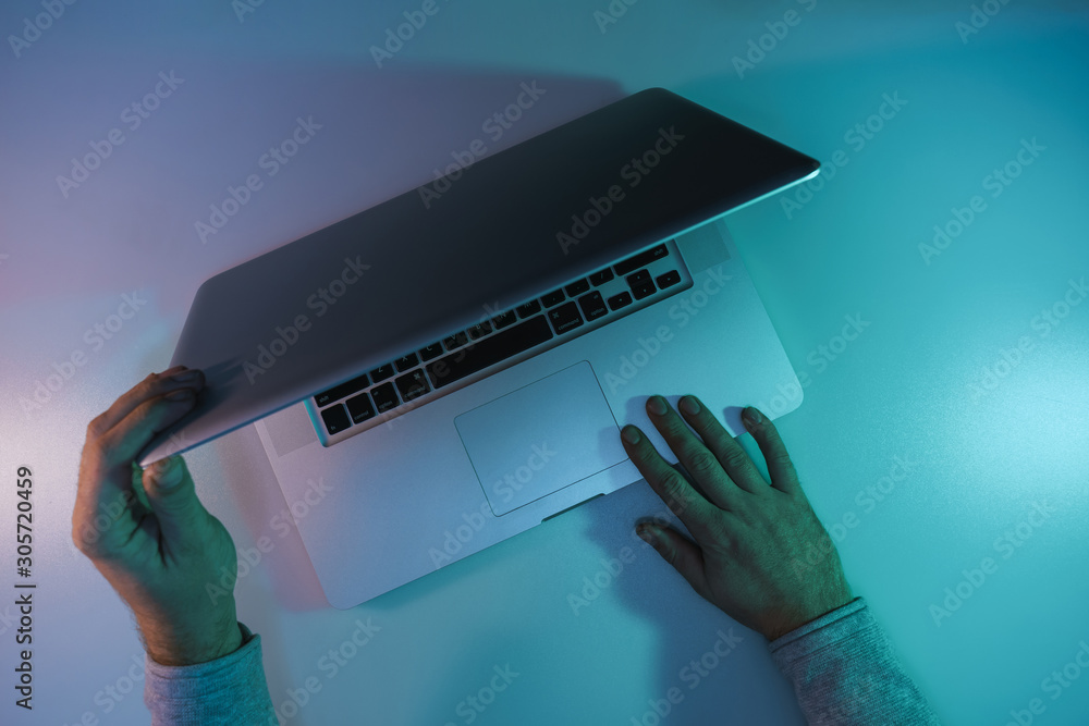 Aufkleber Man open silver laptop night online in the color light on a table night background. Top view.