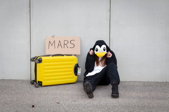 Young woman, with penguin costume and yellow suitcase, has to travel due to global warming. Symbolizes a sad penguin who has to leave his homeland because of global warming or climate change.
