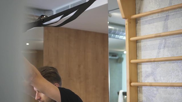 Waist uo view of good-loomung, confident brunet hair man looking aside. He working out with TRX gym equipment in light sport club interior after long day. Making hard exercises at cross fit room