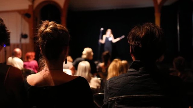 View from the back of audience applauding female speaker in a theater as she leaves the stage with microphone. Slow motion shot with selective focus
