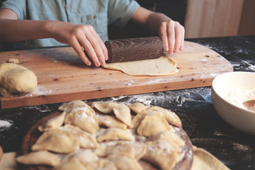  Boy makes dumplings or ravioli from the dough in the kitchen at home. Child cooking concept. On the dark table: child's hands, flour, wooden chopping board. Top view.        