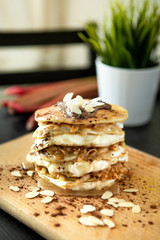 Stack of delicious pancakes with chocolate, mascarpone cream, almonds and apples, topped by honey, laid on a wooden base. Plant in the background.