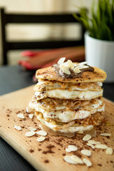 Stack of delicious pancakes with chocolate, mascarpone cream, almonds and apples, topped by honey, laid on a wooden base. Plant in the background.