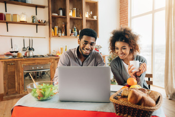Happy african-american couple using laptop at kitchen - Powered by Adobe