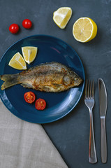 Fried appetizing fish crucian carp with seasoning on a blue plate on a gray background decorated with slices of tomato and lemon with a knife and fork. vertical frame.