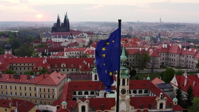 Aerial view through waving flag on foreground. Lateral movement, drone shot, flying European union flag in front of red tiled roofs and famous Prague Castle on horizon. Scenic panorama over Prague