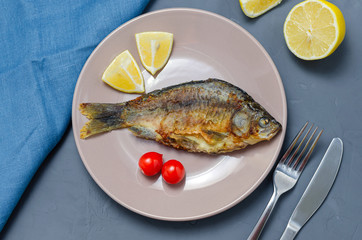 Fried appetizing fish crucian carp with seasoning on a beige plate on a gray background decorated with slices of tomato and lemon with a knife and fork. healthy eating concept.