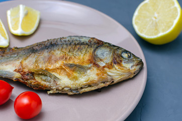 Fried appetizing fish crucian carp with seasoning on a beige plate on a gray background decorated with slices of tomato and lemon. Mediterranean diet. healthy eating concept.