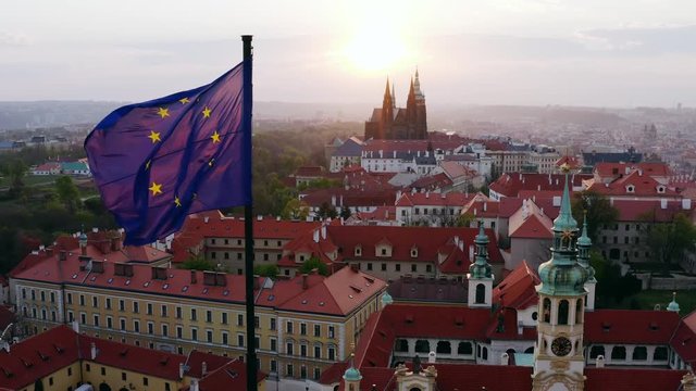 Aerial view town through waving flag on foreground. Lateral movement, drone shot, flying European union flag in front of red tiled roofs at sunrise. Scenic panorama over Prague in loreta district 