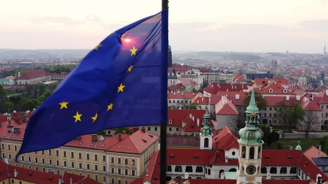 Aerial view town through waving flag on foreground. Lateral movement, drone shot, flying flag of EU in front of shining sun over scenic panorama Prague with red tiled roofs at sunrise