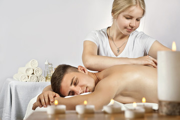 Fototapeta na wymiar Girl in a T-shirt doing a massage to a guy. Candles in the foreground. Man lying on the table on a white background.