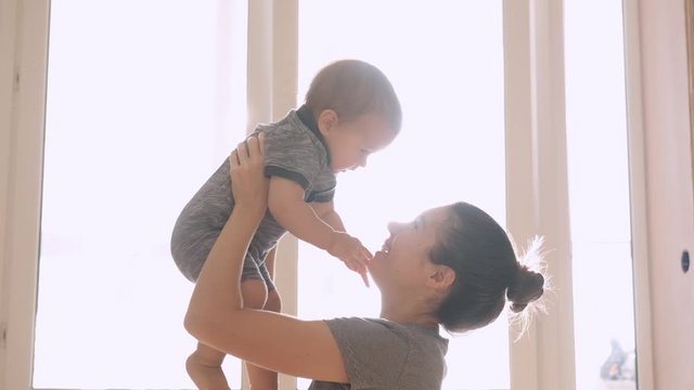 Mother and her baby son having fun and playing at home. Little kid 1 years old play with his mom arms at home near a big window, slow motion. They feel happy, smile and laugh