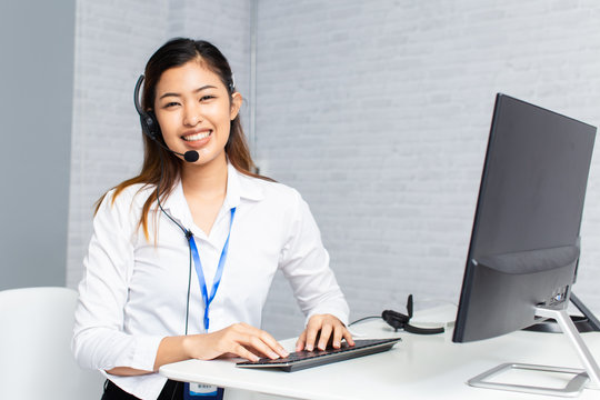 Happy Asian woman with headset smiling and looking at camera while using computer during work in office of call center