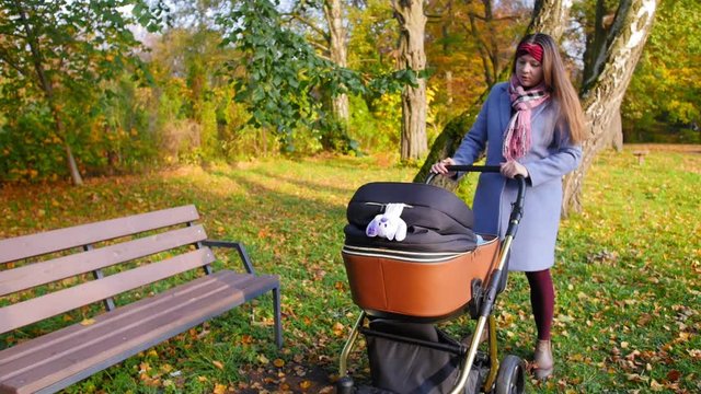 Young happy mother stands with a stroller in the park, smiles at the baby and takes the baby out of the stroller. Full shot.