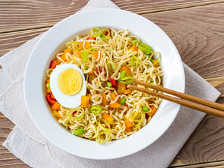 Close-up of instant noodles with egg and vegetables in white bowl and chopsticks on a brown wooden table. Convenience fast food.