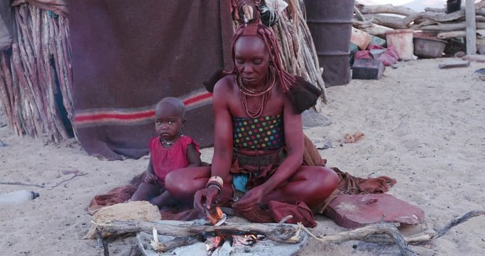 4K side view of Himba woman in traditional dress with young child, filling a pipe and smoking outside their hut within their small compound, Namibia