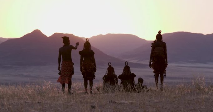 4K view of people from the Himba tribe in traditional dress, watching the sun setting on the mountains, Namib desert, Namibia