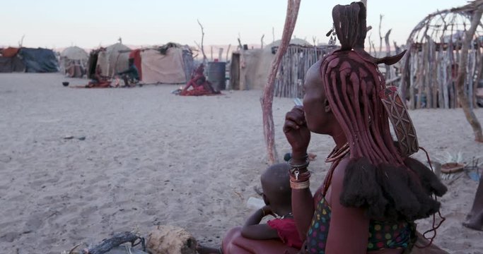 4K side view of Himba woman in traditional dress with young child, sitting and smoking outside their hut within their small compound, Namibia