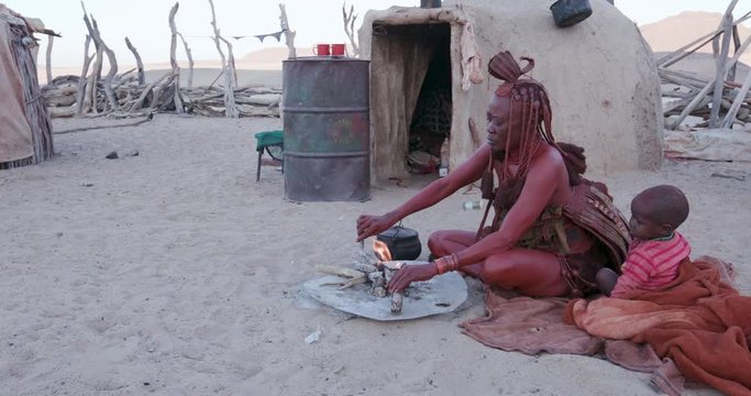 4K view of Himba woman in traditional dress with young child, putting a small pot on a fire outside their hut within their small compound, Namibia