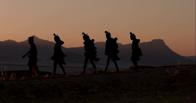 4K view of people from the Himba tribe in traditional dress, walking along a path at sunset with scenic mountains in the background, Namib desert, Namibia