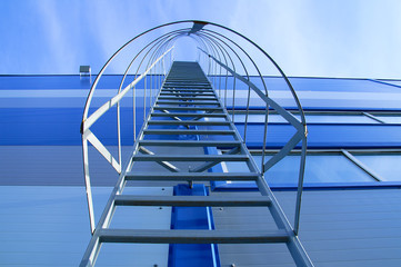 A metal staircase on the facade of an industrial blue building. View from below.