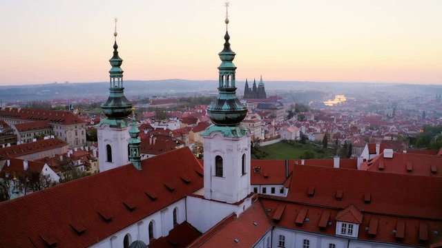 Aerial view over Strahov monastery at sunrise. Lateral shot, drone flying opposite belfries of cathedral decorated crucifix on spire, showing picturesque panorama above Prague in early morning