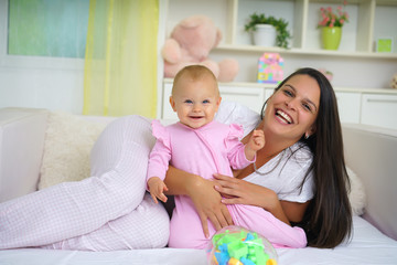  Happy mother and baby girl playing with a baby toys in the bed, looking at camera 