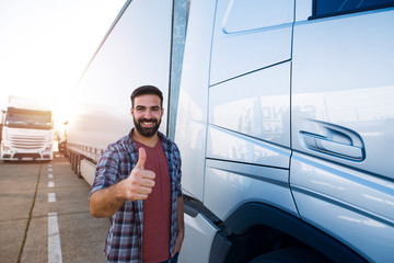 Portrait of young bearded man with thumbs up standing by his truck. Professional and positive truck driver standing by semi truck vehicle. Transportation services. - obrazy, fototapety, plakaty
