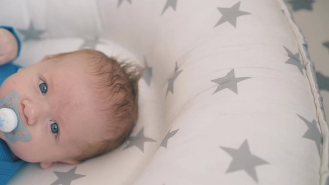 cute newborn boy with large blue eyes sucks pacifier resting in soft sleeping cocoon in cot extreme close view