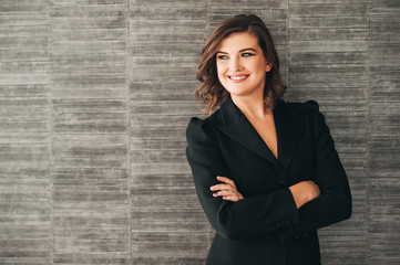 Interior portrait of young successful woman, wearing black suit, posing by the grey brick wall, arms crossed