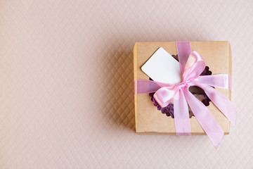 A square gift box with a pink ribbon on a light pink background. Cupcakes inside. Space for text, empty note tied over.